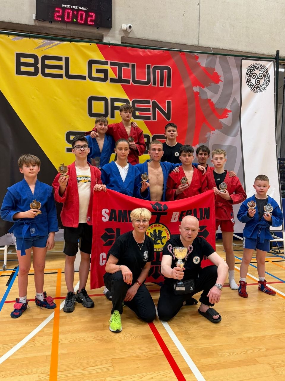 Group of young judo/karate athletes posing with trophies in a gym, yellow Belgium Open banner behind them, red and blue uniforms present.