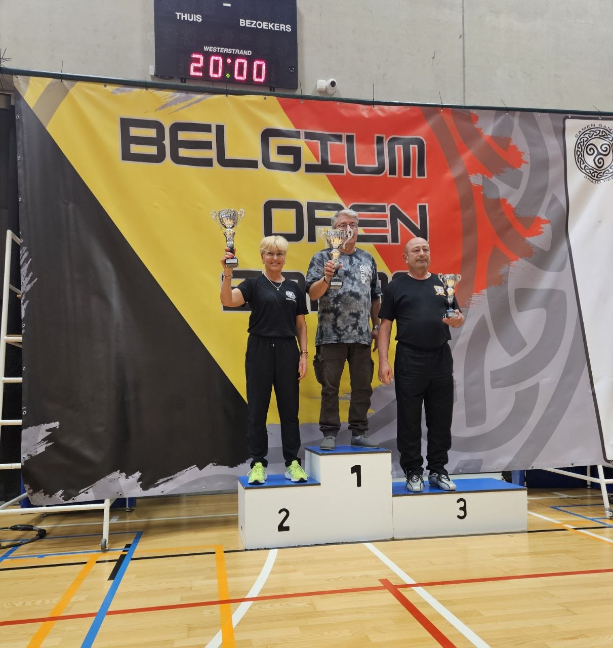 Three podium finishers hold trophies during a Belgium Open ceremony on an indoor gym floor, with a bold yellow-black banner behind them.
