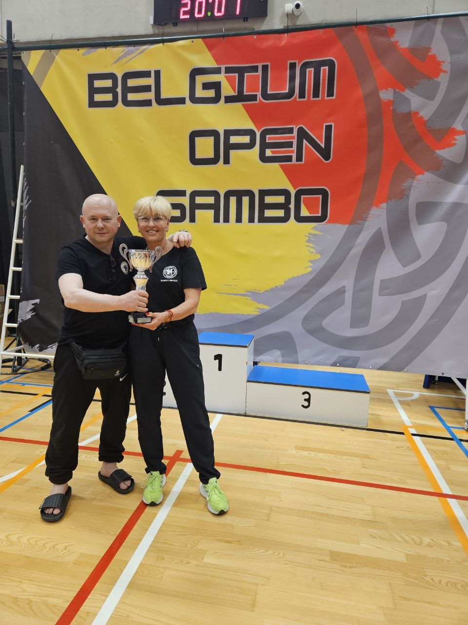 Two people holding a trophy in a gym, standing in front of a Belgium Open Sambo banner and a podium labeled 1 and 3.