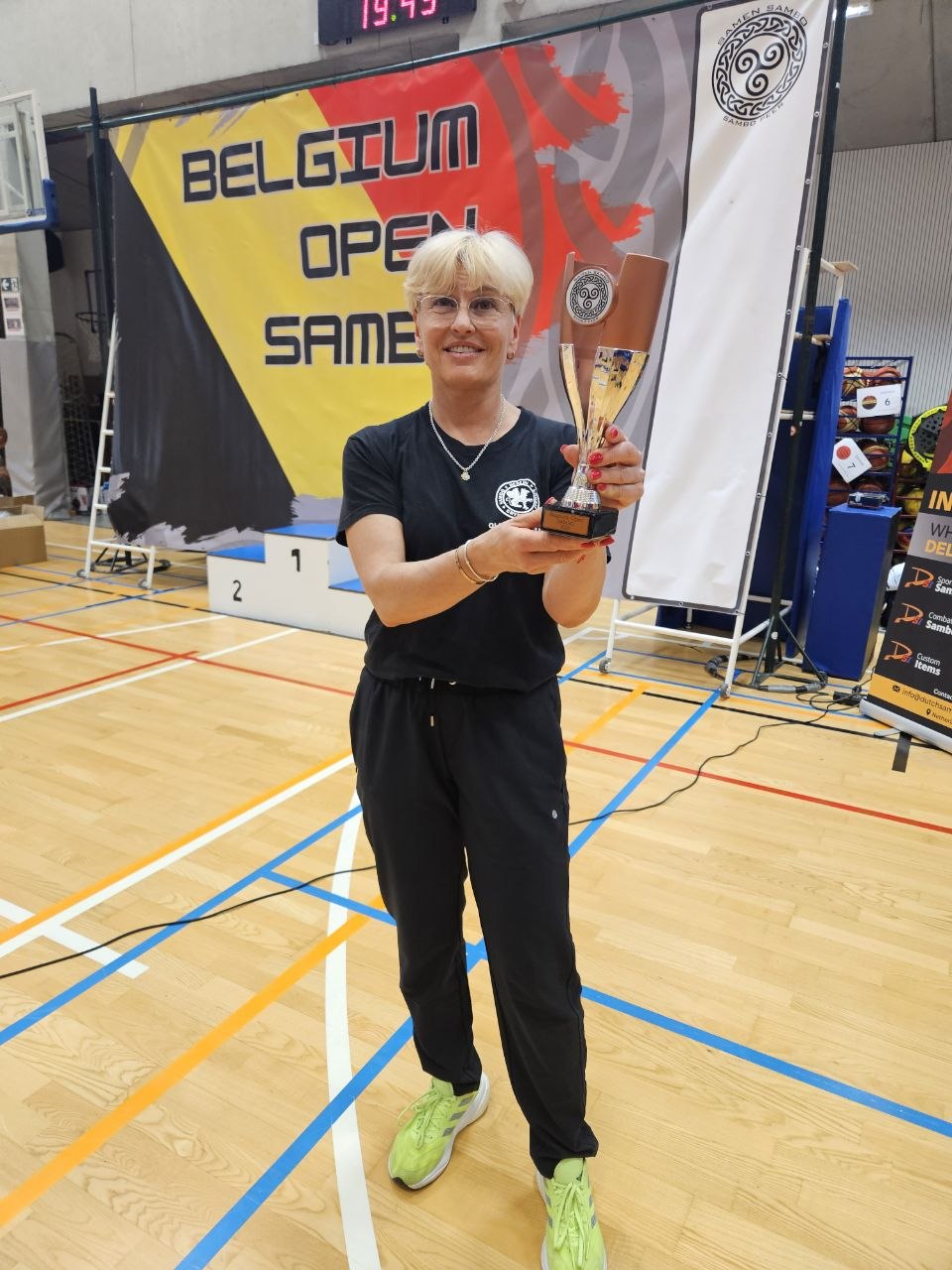 Smiling woman in black sportswear holding a trophy inside a gym with a Belgium Open Sambo banner in the background.