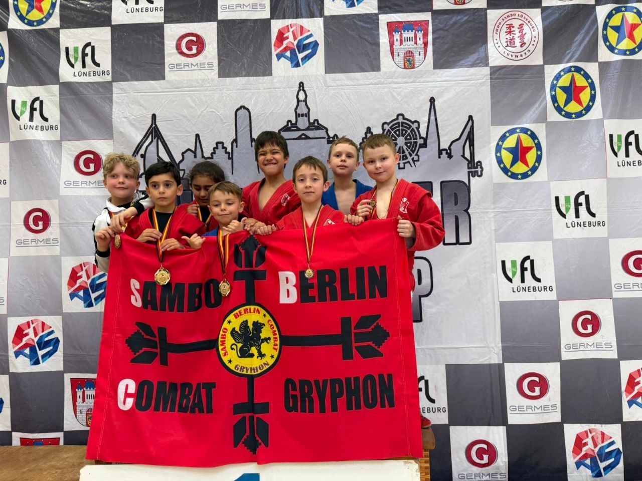 Group of smiling kids in red uniforms posing with medals behind a red banner that reads 'SAMBO BERLIN COMBAT GRYPHON' at a sponsor backdrop.