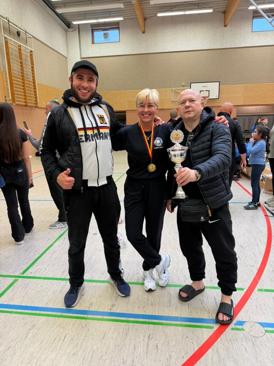 Three people pose in a gym after a competition: a man in a cap, a smiling woman with a gold medal, and a man holding a large trophy.