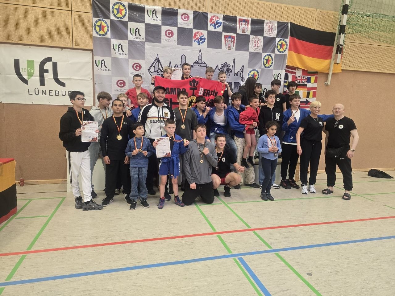 Group of children and teens with coaches posing for a photo after a martial arts competition, medals and certificates visible behind them in a gym setting.