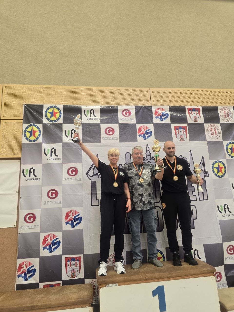 Three winners stand on a first-place podium, holding trophies and wearing medals, with a sponsor banner in the background.
