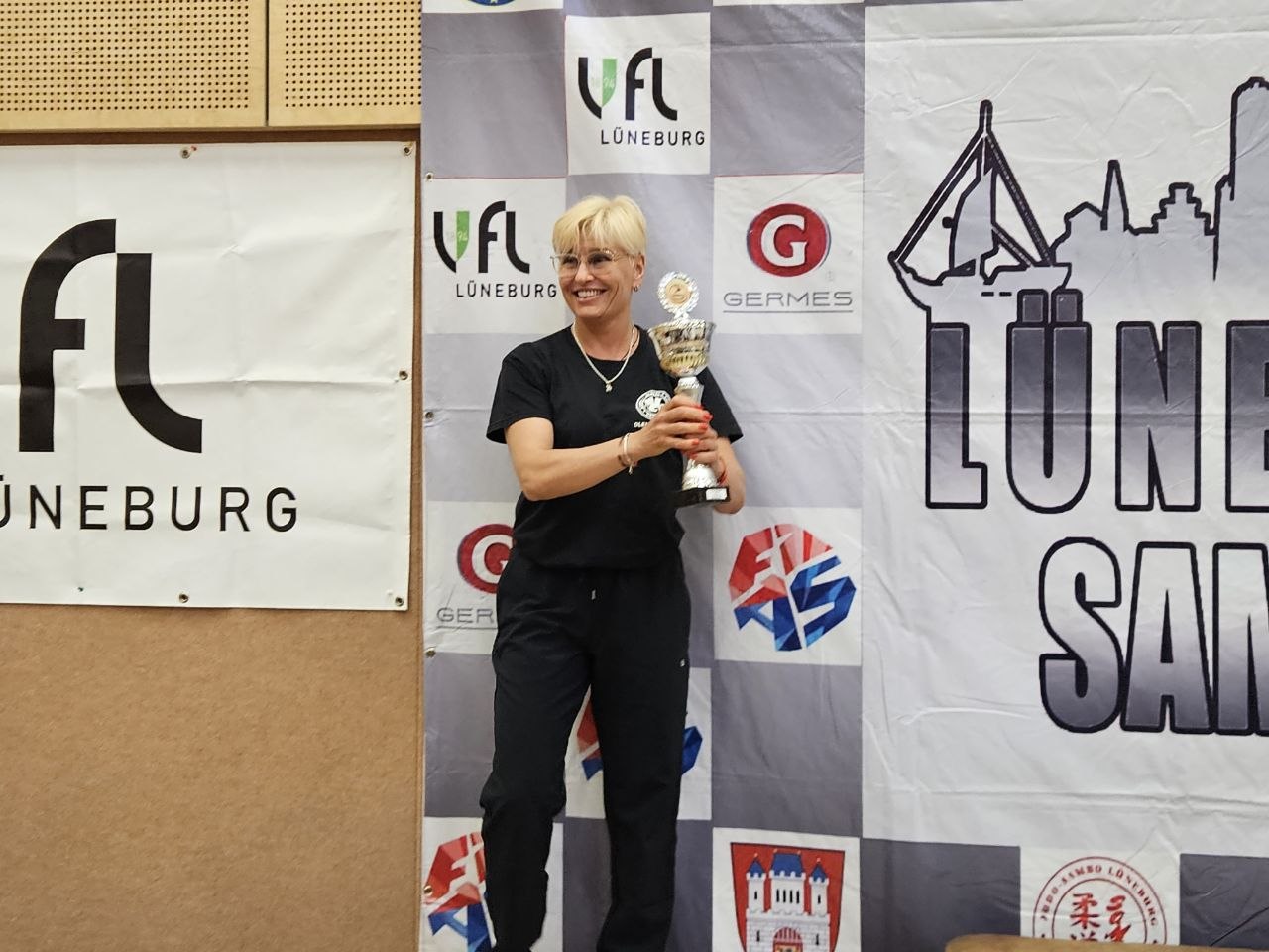 Woman on a stage holding a trophy, smiling, with a banner of logos and cityscape behind her in an award ceremony setting.
